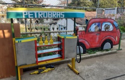 Maquete colorida de posto de gasolina, com desenho de carro ao lado, durante o dia.