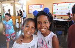 Duas meninas sorriem; ao fundo, à esquerda, crianças jogam pebolim. 