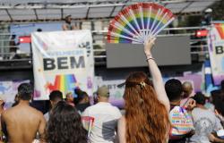 Pessoas de costas para o palco participam da Parada LGBT. Em destaque, uma mulher branca, ruiva, segura um leque nas mãos