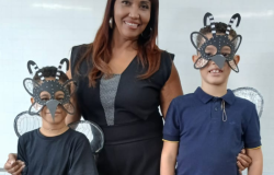 #paratodosverem: a foto mostra uma professora entre dois estudantes. As crianças usam máscaras e asas representando o mosquito da dengue. Todos(as) estão sorrindo. O fundo é um quadro branco e parte de uma parede de azulejos brancos. 