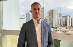Diretor de Negócios - Daniel Rodrigues Nogueira, posa em uma fotografia em local fechado com palito tom azul claro, camisa cor branca ao lado de uma janela com vários prédios.