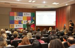Auditório com grande público sentado em cadeiras. pessoas acompanham atentamente uma apresentação. O ambiente possui piso de madeira, cortinas marrons nas laterais e paredes revestidas com painéis acústicos vermelhos. A maioria dos participantes usa crachás de identificação. As pessoas estão voltadas para a parte da frente do auditório