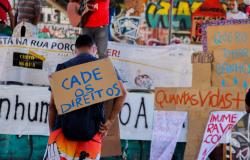 A fotografia mostra uma manifestação em defesa dos direitos da população em situação de rua. Em primeiro plano, uma pessoa de costas, com cabelos curtos, camiseta azul-escura, mochila preta e bermuda colorida, carrega nas costas um cartaz de papelão com a frase “Cadê os direitos?”, escrita à mão em letras azuis. Ao fundo, aparecem outras pessoas, faixas e cartazes com reivindicações, incluindo um com a frase “Quero tomar banho”. Uma pessoa segura um violão. O espaço é marcado por paredes cobertas de grafite