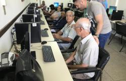 Dois idosos, sentados cada um em frente ao um computador, recebem orientações de monitor, que está de pé. 