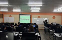 Sala de aula, com computadores nas mesas, alunos presentes e professor em frente ao quadro branco.