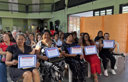 Uma sala cheia de idosos, na maioria mulheres, brancas, pardas e pretas, seguram o certificado de conclusão do curso.