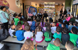 #paratodosverem: na foto, crianças sentadas em fileiras de bancos assistem a uma apresentação musical realizada pela banda da Guarda Municipal. Eles tocam instrumentos e cantam em frente a um mural decorado com desenhos e mensagens. Um banner com o brasão da escola aparece ao fundo, junto a outras pessoas adultas sentadas e em pé, que acompanham e aplaudem a atividade.