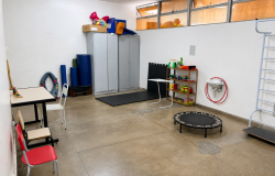 Sala equipada com materiais de reabilitação. Um armário no fundo da sala, uma mesa com cadeiras na lateral esquerda, um trampolim, tatames e brinquedos na lateral direita. 