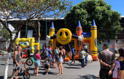  Rua fechada para atividades de lazer, em um dia de céu limpo e ensolarado. Ao centro, brinquedos infláveis recebem crianças, enquanto adultos permanecem ao redor acompanhando a movimentação. Árvores de grande porte fazem sombra sobre parte da via. Ao fundo, há um prédio e um muro com pinturas, compondo um ambiente de convivência, recreação e encontro entre famílias.
