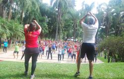 Dois instrutores orientam mais de trinta pessoas a fazer ginástica no parque, durante o dia. 