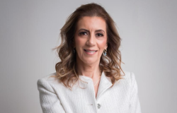 Mulher branca, cabelos castanhos, ondulados e longos. Usa blazer branco.