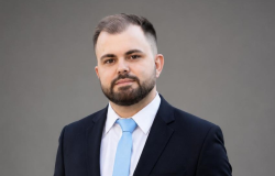 Homem jovem, de pele clara, com barba e cabelo curtos, usando terno escuro, camisa branca e gravata azul-clara, olhando para a câmera com expressão séria, diante de fundo cinza.
