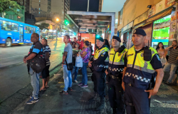 Três guardas municipais ao lado de um ponto de ônibus com mais de seis pessoas, à noite. 