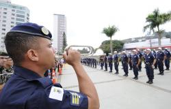 Guarda civil municipal toca instrumento de sopro; à frente, mais de trinta guardas enfileirados. 