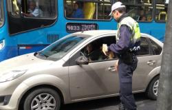 Guarda municipal entrega folder educativo a motorista de carro particular. Um ônibus aparece ao fundo da imagem