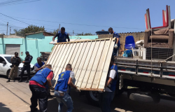 Três homens com colete azul colocam portão de ferro em caminhão, durante o dia. 