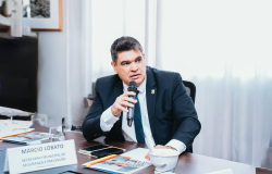 Homem sentado à mesa, usando terno escuro, camisa branca e gravata azul, segurando um microfone enquanto fala. Ao fundo, há uma janela com cortinas claras e, sobre a mesa, estão documentos, celulares, um copo e uma pequena placa com a identificação “Márcio Lobato — Secretário Municipal de Segurança e Prevenção”