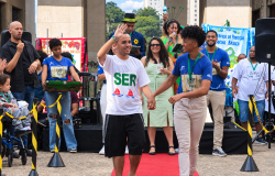 Fotografia colorida de um evento ao ar livre, realizado em uma área pavimentada, com prédios e árvores ao fundo. Ao centro da imagem, um jovem caminha por um tapete vermelho. Ele usa camiseta branca com a palavra “SER” escrita em letras verdes e bermuda preta. Com uma das mãos, faz um gesto de aceno e, com a outra, segura a mão de uma pessoa que caminha ao seu lado.