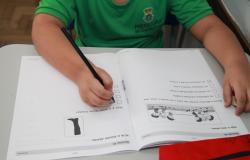 Estudante com camiseta verde da Prefeitura de Belo Horizonte realiza atividade em um caderno aberto sobre a mesa, utilizando um lápis preto para responder às questões. Na página, há exercícios com figuras e alternativas para marcar.