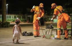 Criança observa dois gari varrendo uma praça à noite.
