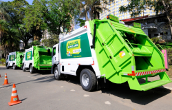Três caminhões de coleta seletiva nas cores verde e branco, na rua, enfileirados, durante o dia. 