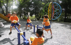 Quatro alunos da Rede Municipal de Educação, com camisetas laranja, brincam em brinquedos de uma praça, durante o dia. 