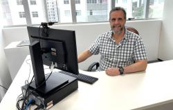 Homem sentado em frente ao computador, usando camisa xadrez, posa para a foto