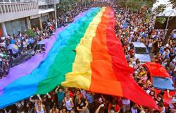 Mais de 800 pessoas em uma rua com uma imensa faixa com as cores do arco-íris, símbolo da luta LGBT, durante o dia.