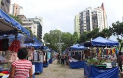 Barraquinhas de artesanato azuis, enfileiradas, com pessoas visitando, durante o dia. Ao fundo, prédios. 