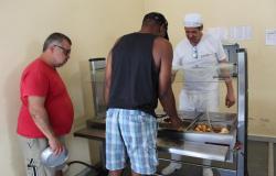 Homem de branco e touca serve comida para dois homens de bermuda. 