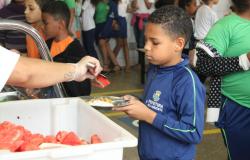 Foto de criança usando uniforme da Prefeitura de Belo Horizonte segurando um prato de comida. Há uma pessoa servindo uma melancia.