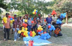 Grupo de vinte pessoas posa para a foto em um jardim. Eles seguram balões nas cores azul, amarelo e laranja