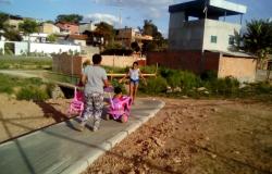 Duas mulheres andam em passeio recém feito; uma delas empurra carrinho rosa com criança e carrega outro carrinho vazio nas mãos. 