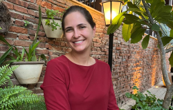 Mulher adulta, de cabelos escuros presos, pele clara e olhos castanhos. Apresenta um sorriso simpático e veste uma blusa de manga longa na cor vinho. Está em um ambiente externo com plantas e parede de tijolos aparentes.
