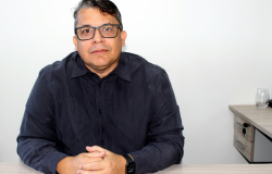 Homem de meia-idade, de pele clara, com cabelos curtos escuros e fios grisalhos nas laterais. Usa óculos de armação preta, camisa social de manga longa em tom escuro, relógio inteligente no pulso esquerdo e aliança. Está sentado à mesa com as mãos entrelaçadas, olhando diretamente para a câmera, com expressão neutra e postura formal.