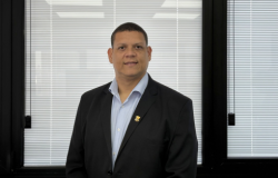 Homem de pele morena, cabelos curtos e escuros, olhando para a câmera com leve sorriso. Veste blazer preto sobre camisa social azul-clara, sem gravata, e usa um pequeno broche dourado na lapela. Está em pé, com postura ereta, transmitindo uma aparência formal e profissional.