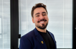 Homem de aparência jovem, com cabelos curtos castanhos, barba e bigode. Está vestido com blazer azul-marinho sobre camiseta preta, usando um broche na lapela, em um ambiente corporativo moderno e bem iluminado. A imagem passa uma impressão de credibilidade, acessibilidade e profissionalismo.