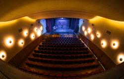 Imagem do teatro. Acentos no meio, palco ao fundo iluminado e com as cortinas abertas, luzes redondas nos corredores laterais. 