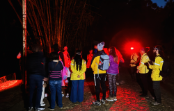 Pessoas reunidas de costas para a câmera, todas vestidas com calças, blusas de frio, tênis esportivo e carregando bolsas e mochilas. A foto foi realizada no período da noite, em local aberto e com muita natureza ao redor. Uma das pessoas carrega uma lanterna de luz vermelha que ilumina toda a região. 
