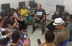 Em uma sala um grupo de pessoas sentam em cadeiras em roda, algumas conversando entre si e outras usando o celular. Atrás das pessoas existem computadores em mesas. 