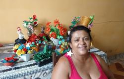 Mulher com chapéu de artesanato, à frente de mesa com arranjos de flores artificiais e bonecas decoradas. 