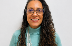 Mulher de cabelos longos, escuros e cacheados, usando óculos de armação escura. Está sorrindo, veste uma blusa de gola alta verde-claro e um colar longo branco, em frente a um fundo claro e neutro.