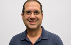 Homem de aparência simpática e sorridente, com cabelo curto e escuro, óculos de armação fina e barba. Veste uma camisa polo azul-marinho e está posicionado em frente a um fundo claro, transmitindo uma imagem profissional e acolhedora.
