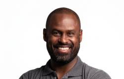 Homem negro, adulto, com barba curta e cabelo bem baixo, sorrindo de forma simpática. Ele veste uma camisa polo cinza e está com os braços cruzados, em frente a um fundo branco.