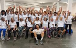 Participantes do Programa Vida Ativa, da Prefeitura de Belo Horizonte, reunidas em um salão comunitário. Vestindo camisetas brancas do programa, as mulheres aparecem sorrindo e com os braços levantados, ao lado de um professor de Educação Física sentado à frente do grupo. A imagem registra um momento de integração, alegria e convivência entre as participantes.