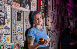 Fotografia em plano médio de uma mulher jovem, parda, com cabelos cobertos por um lenço azul ornamentado e diversas tatuagens no pescoço e braços. Ela veste uma camiseta azul e segura um pequeno prato com bolinhos fritos empanados e dourados.