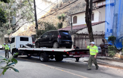 Um caminhão-guincho branco está parado em uma rua íngreme e gramada para rebocar um carro preto danificado em uma área residencial arborizada. Dois agentes da BHTrans coordenam a operação, com um deles sinalizando na frente do veículo e o outro posicionado na parte traseira.