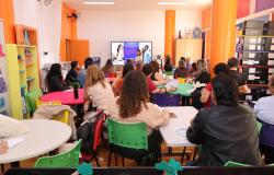 A foto mostra uma sala ampla e colorida, com várias mesas redondas e cadeiras de diferentes cores. Cerca de 20 pessoas estão reunidas, aparentemente participando de uma palestra ou treinamento, voltadas para uma televisão na parte da frente da sala. A tela exibe uma apresentação com imagens e textos. O ambiente possui paredes e colunas em tons de laranja, armários, estantes com materiais organizados e ventiladores nas paredes. A cena transmite um clima de reunião, aprendizado e interação.