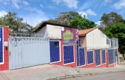 Fachada da Escola Municipal Lídia Angélica. O imóvel é térreo, com paredes revestidas por pastilhas nas cores azul, vermelho e branco e portões metálicos. A entrada e os muros são protegidos por concertina. Na fachada há uma placa de identificação e a logomarca da Prefeitura. O prédio possui telhados de diferentes alturas, janelas com grades e uma área de acesso com rampa de concreto. Em frente à escola, há uma calçada e a rua. Ao fundo, tem árvores de grande porte sob. céu azul com nuvens