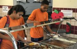 Dois alunos adolescentes , um menino e uma menina, usando camisetas cor de laranja estão em frente à um bandejão de uma cantina, servindo seus pratos.